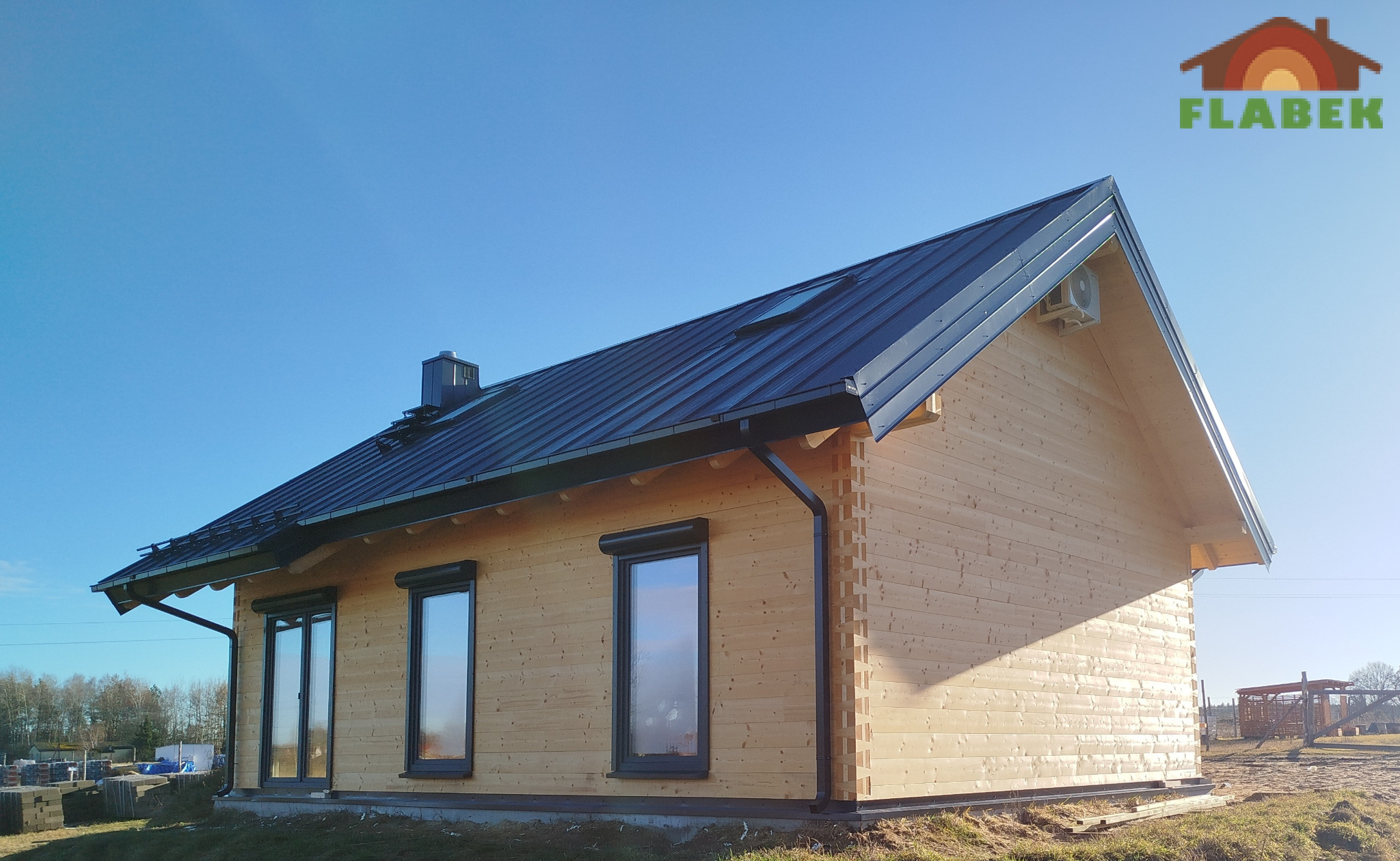 Jasny, parterowy dom z bali z ciemnym dachem i roletami zewnętrznymi, widziany w słoneczny dzień. Widoczny komin i klimatyzator na ścianie. Logo firmy w prawym górnym rogu.