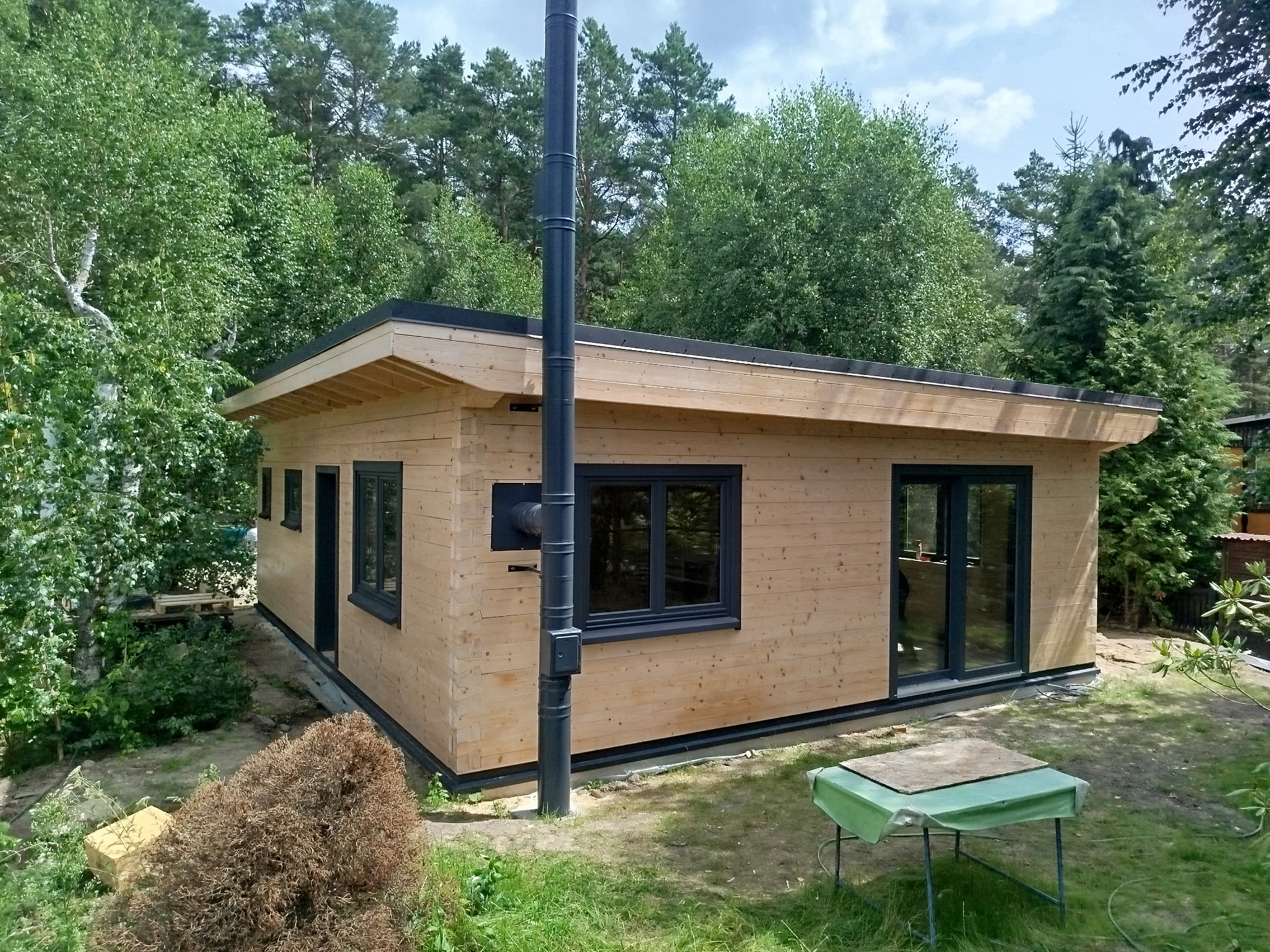 Gotowy, parterowy dom z bali z czarnymi oknami i kominem na tle zieleni. Widok z zewnątrz, prezentujący elewację i otoczenie.