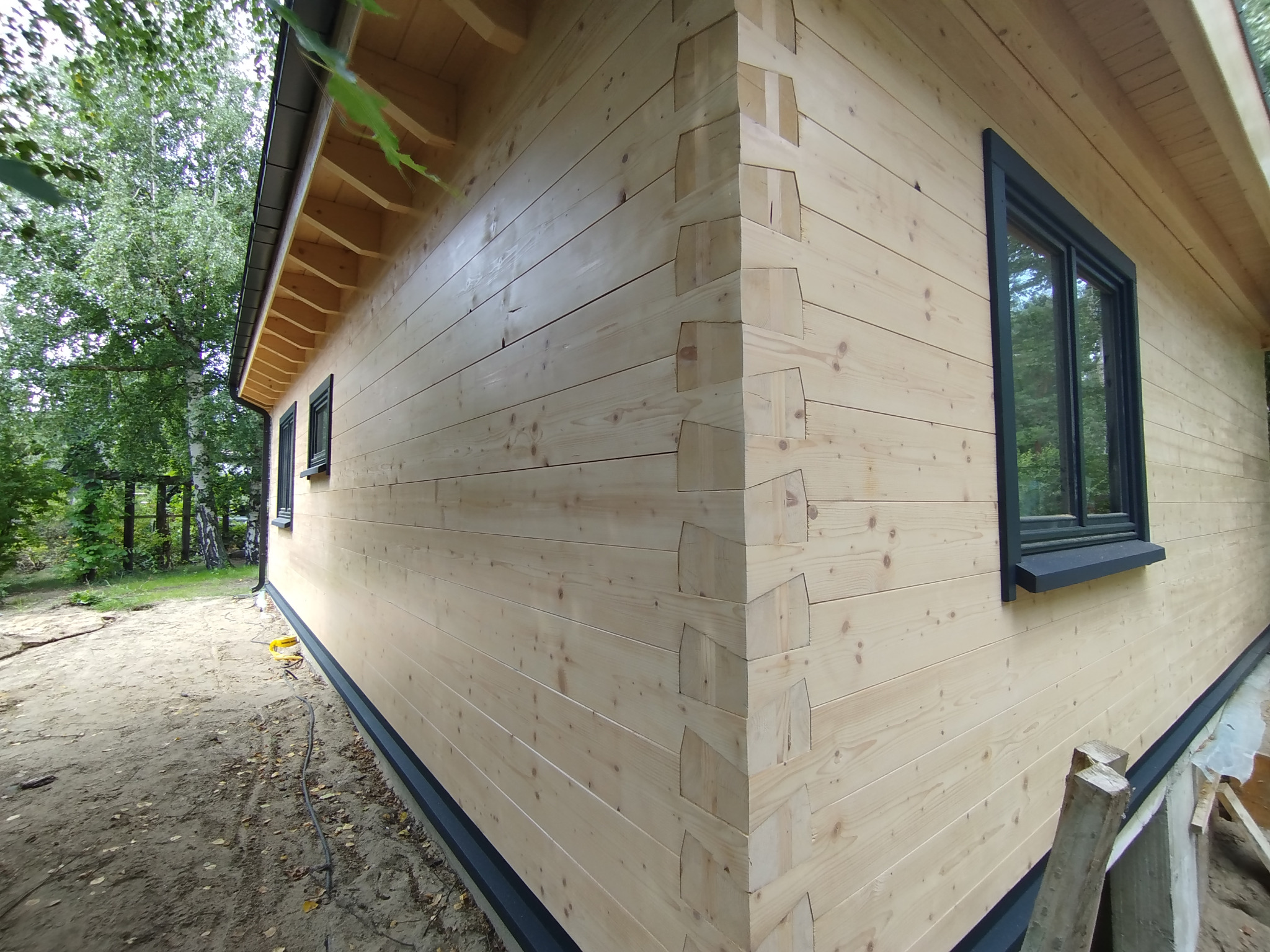 Zewnętrzny róg domu z bali z widocznymi połączeniami na węgłach, czarne ramy okienne, jasne drewno, zielone drzewa w tle, częściowo widoczne narzędzia budowlane.