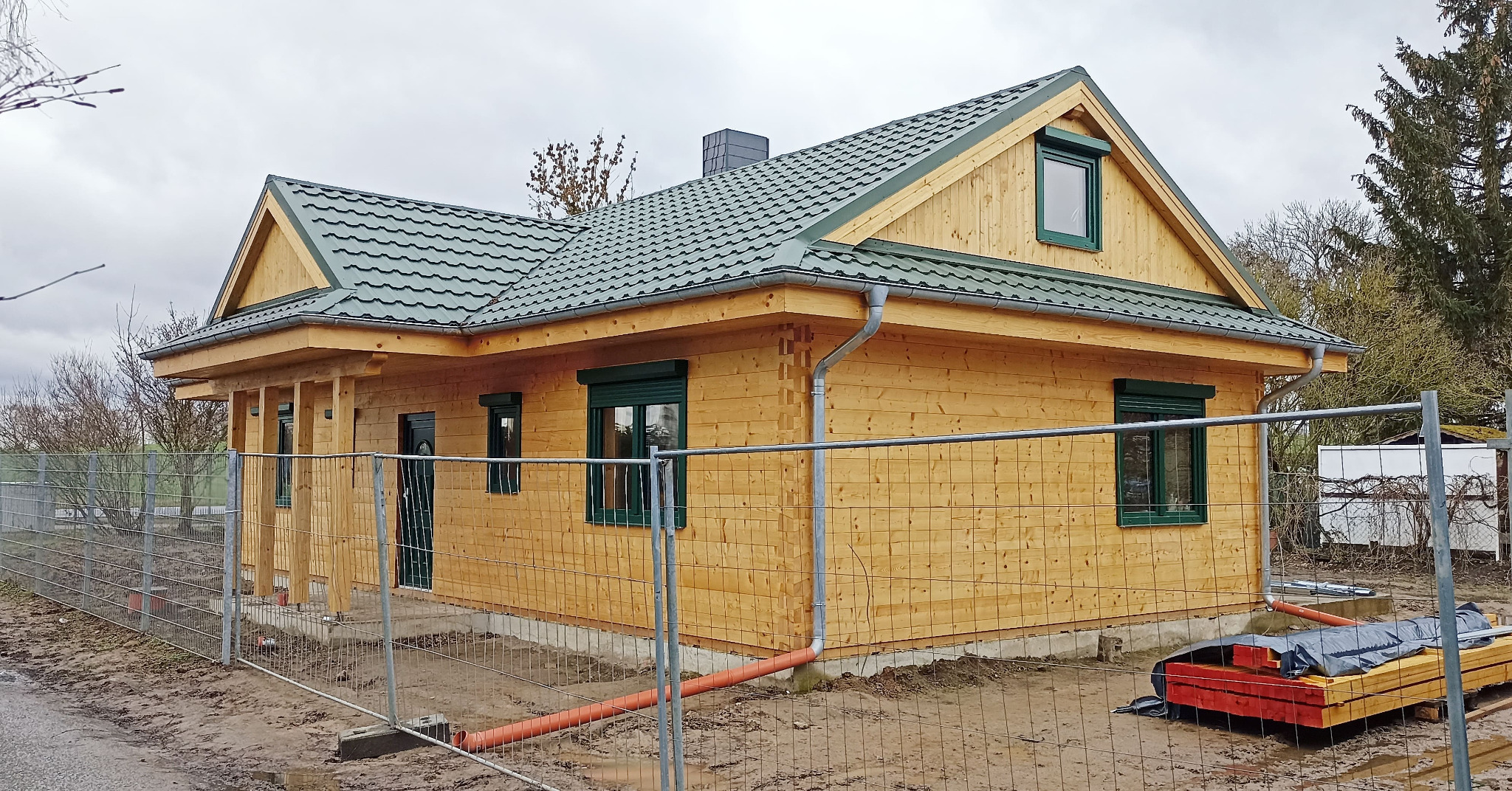 Żółty, parterowy dom z bali z zielonym dachem i oknami, otoczony tymczasowym ogrodzeniem. Widoczne elementy konstrukcyjne i materiały budowlane.