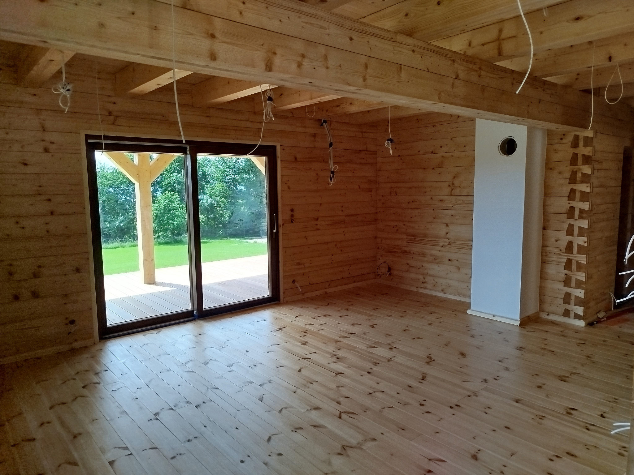 Jasne, drewniane wnętrze domu z bali, z widoczną podłogą, ścianami i sufitem z drewna. Duże przesuwne drzwi balkonowe wpuszczają światło. Widoczne elementy instalacji elektrycznej.
