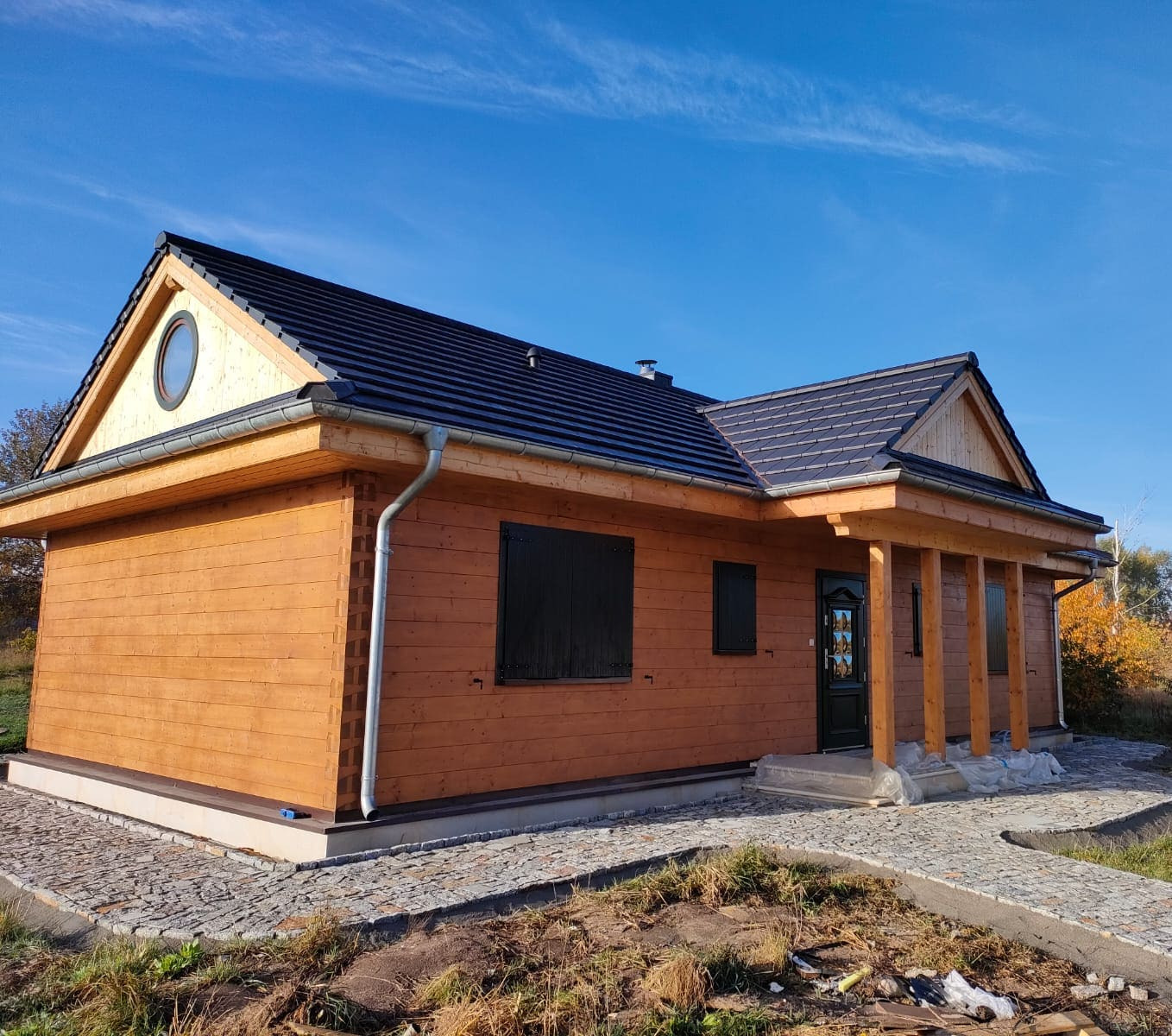 Gotowy, parterowy dom z bali z ciemną dachówką i okrągłym oknem na szczycie, otoczony brukowanym chodnikiem i jesienną roślinnością.
