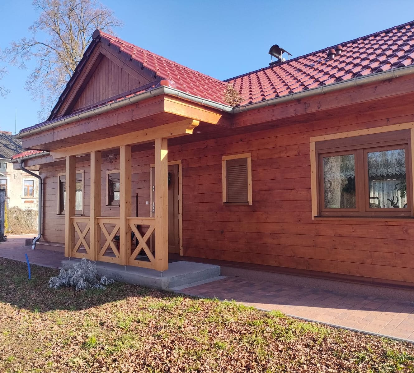 Wykończony dom z bali z czerwonym dachem i drewnianą werandą w Żarach. Widoczne detale wykończenia i otoczenie domu w słoneczny dzień.