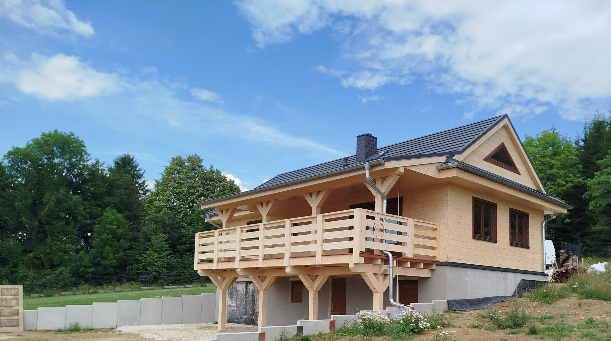 Jasny, drewniany dom z balkonem i ciemnym dachem na tle błękitnego nieba i zieleni drzew. Widoczne elementy wykończeniowe i fragment fundamentów.