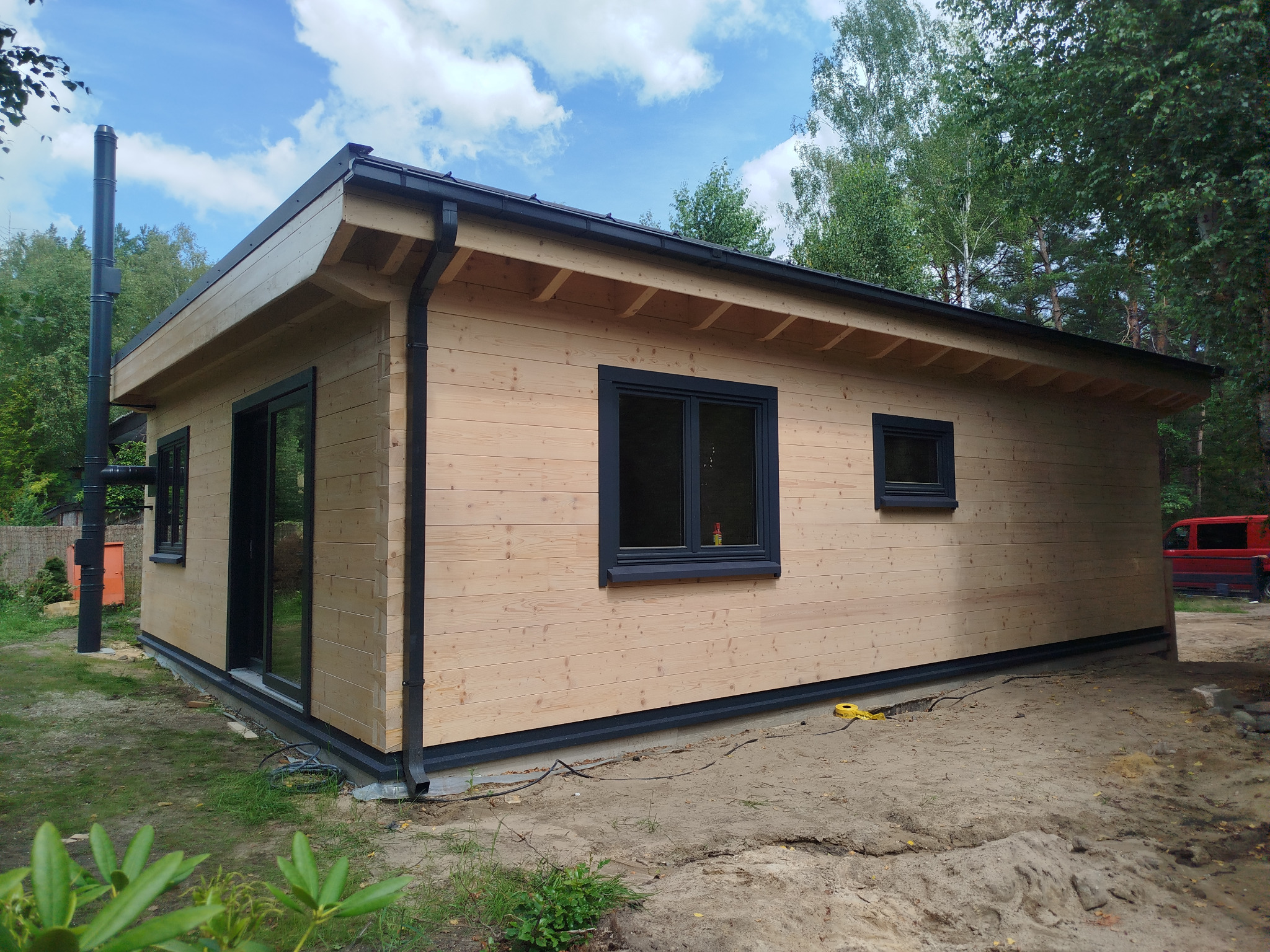 Gotowy, parterowy budynek z jasnego drewna, z czarnymi oknami i rynnami, otoczony zielenią i piaskiem. Widoczny komin i fragment czerwonego samochodu w tle.