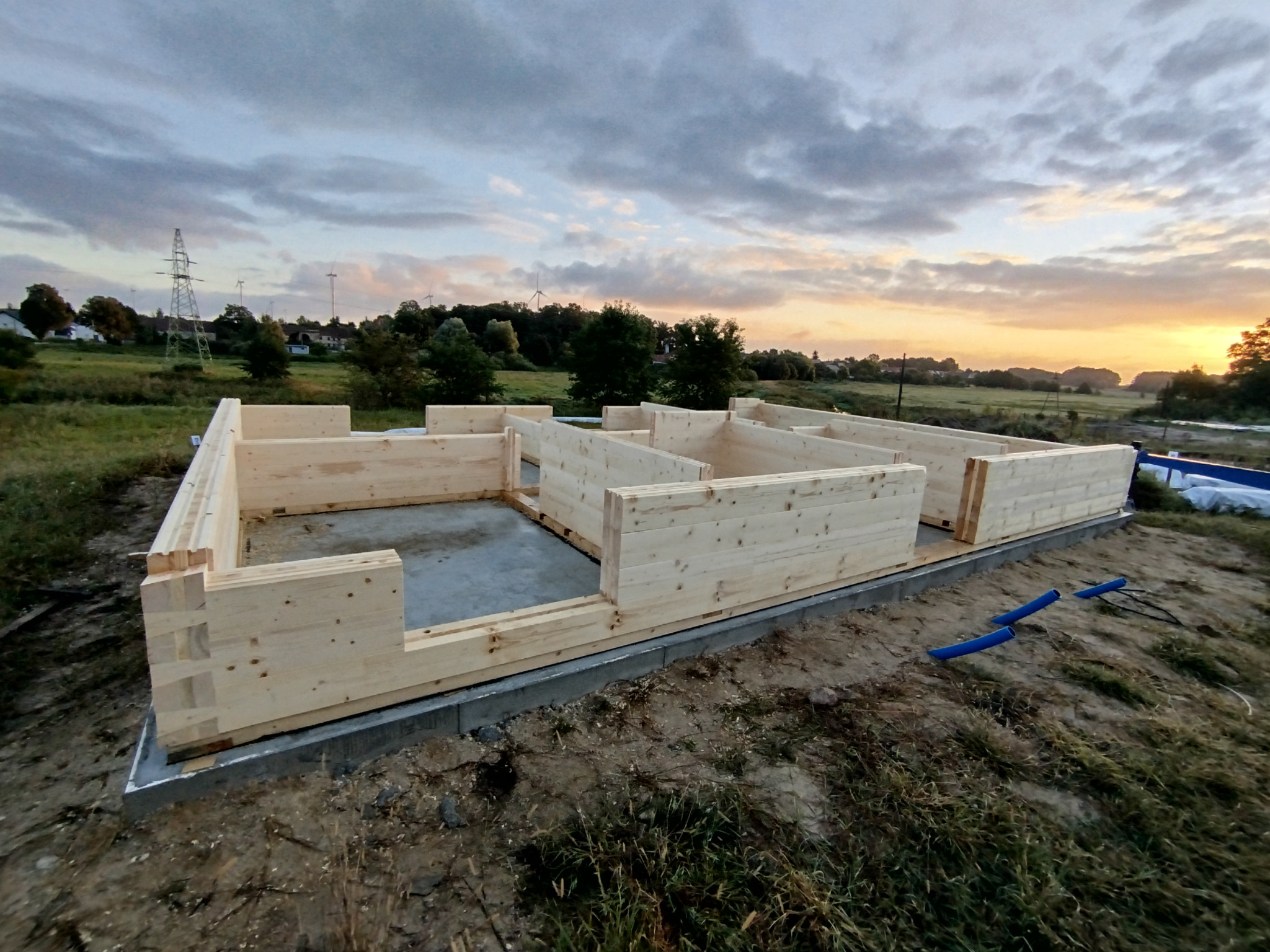 Wznoszenie domu z bali: surowe drewno tworzy ściany na betonowej podstawie, w tle krajobraz z liniami wysokiego napięcia i zachodzące słońce.