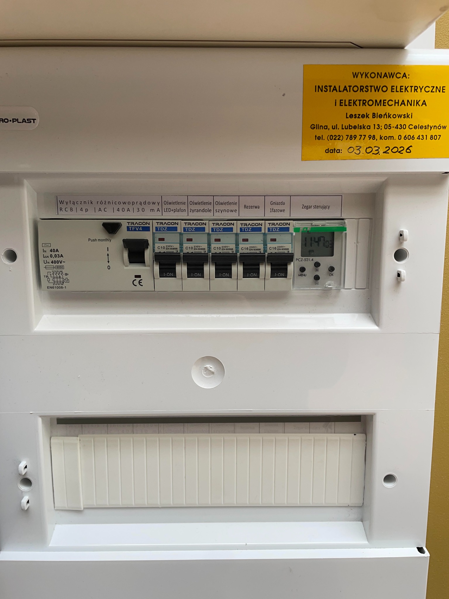 Biała skrzynka rozdzielcza z bezpiecznikami TRACON, wyłącznikiem różnicowoprądowym i zegarem sterującym. Widoczna naklejka z danymi wykonawcy instalacji elektrycznej.