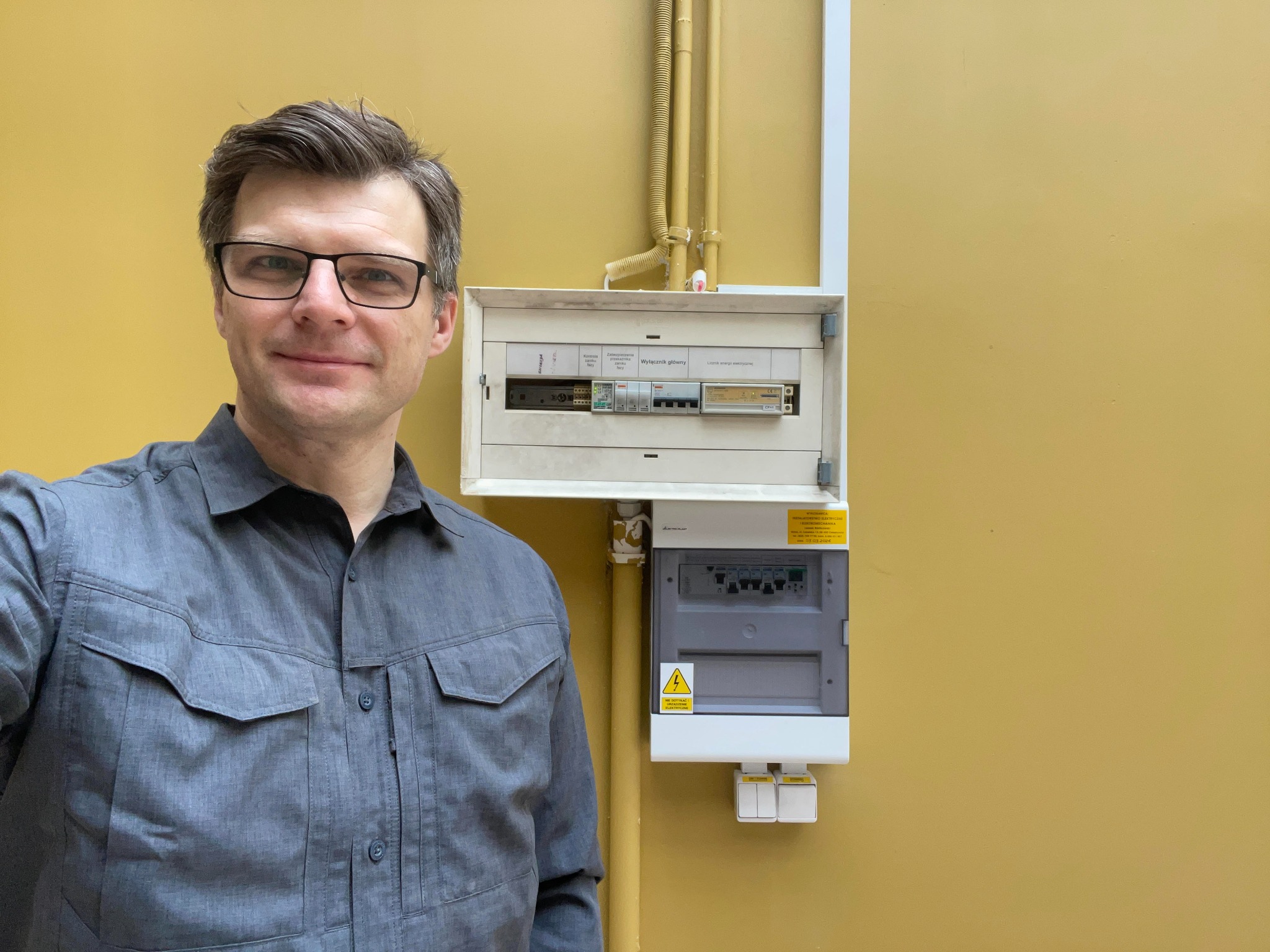 Elektryk prezentuje zamontowaną, dwuczęściową skrzynkę elektryczną na żółtej ścianie. Widoczne przewody w peszlach. Uśmiechnięty mężczyzna w okularach.