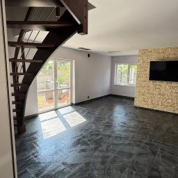 Usługi remontowo budowlane - Salon po remoncie z ciemną, błyszczącą podłogą z płytek, drewnianymi schodami i ścianą z kamienia dekoracyjnego pod telewizorem. Wpadające światło słoneczne.