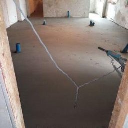 Usługi remontowo budowlane - Wylana, świeża posadzka cementowa w surowym wnętrzu. Widoczne wystające rury instalacyjne i narzędzie do rozprowadzania betonu. Surowe ściany w tle.