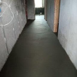 Usługi remontowo budowlane - Wylana, świeża posadzka w wąskim korytarzu z surowymi ścianami. Widoczne kable i niedokończone wykończenie. Minimalistyczny, surowy styl.