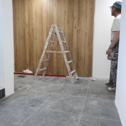 Usługi remontowo budowlane - Montaż paneli ściennych imitujących drewno w pomieszczeniu. Widoczna drabina malarska i fragment sylwetki pracownika. Szare płytki na podłodze.