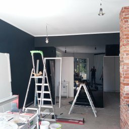 Usługi remontowo budowlane - Wnętrze w trakcie prac wykończeniowych: drabiny, płytki, ściana z cegły i czarna ściana. Widoczny fragment łazienki z umywalką. Osoba robiąca zdjęcie w tle.