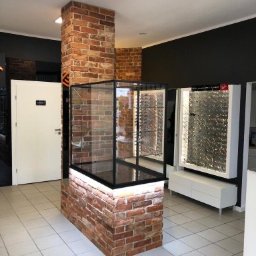 Usługi remontowo budowlane - Wnętrze sklepu optycznego z ceglanym filarem i witryną, oświetleniem LED, białymi meblami i szarą ścianą w tle. Nowoczesny design z elementami industrialnymi.