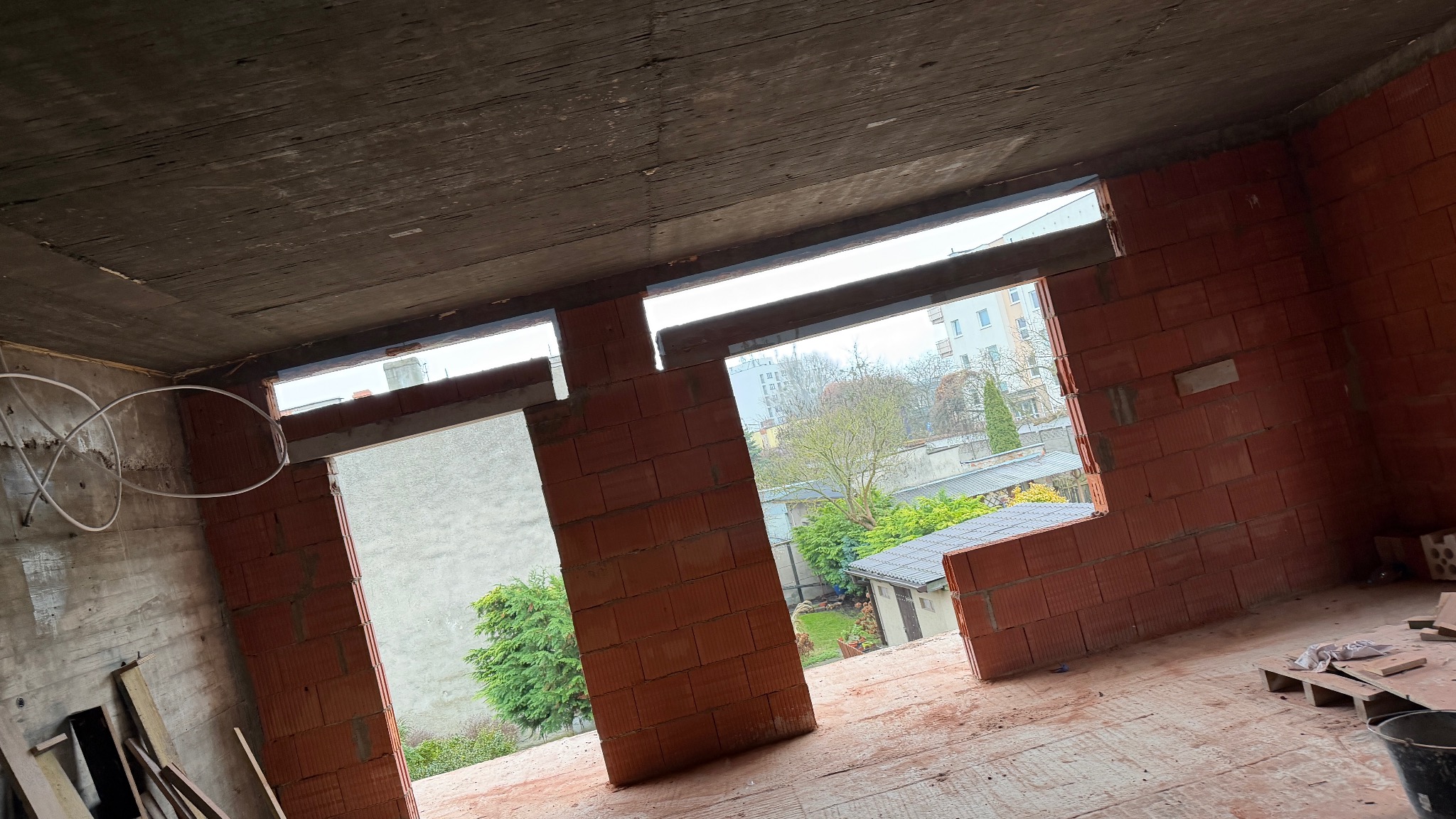 Surowy stan wnętrza budynku z czerwonej cegły, widoczne okna z widokiem na zewnątrz, betonowy sufit i podłoga. W tle zieleń drzew i budynki.