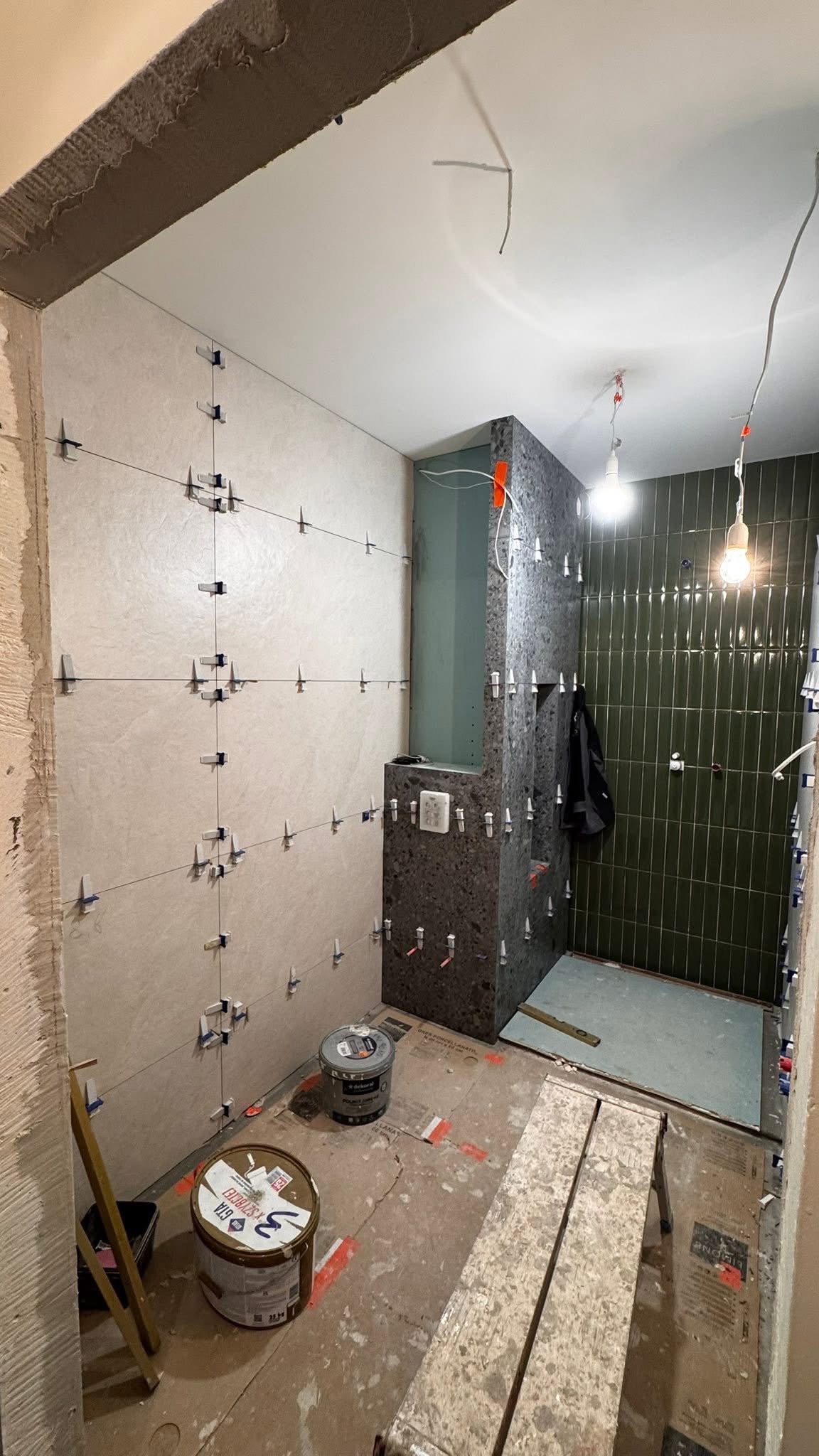 Łazienka w trakcie remontu: jasne i ciemne płytki ułożone z użyciem krzyżyków dystansowych, widoczne wiadra z klejem i drabina. Etap prac wykończeniowych.