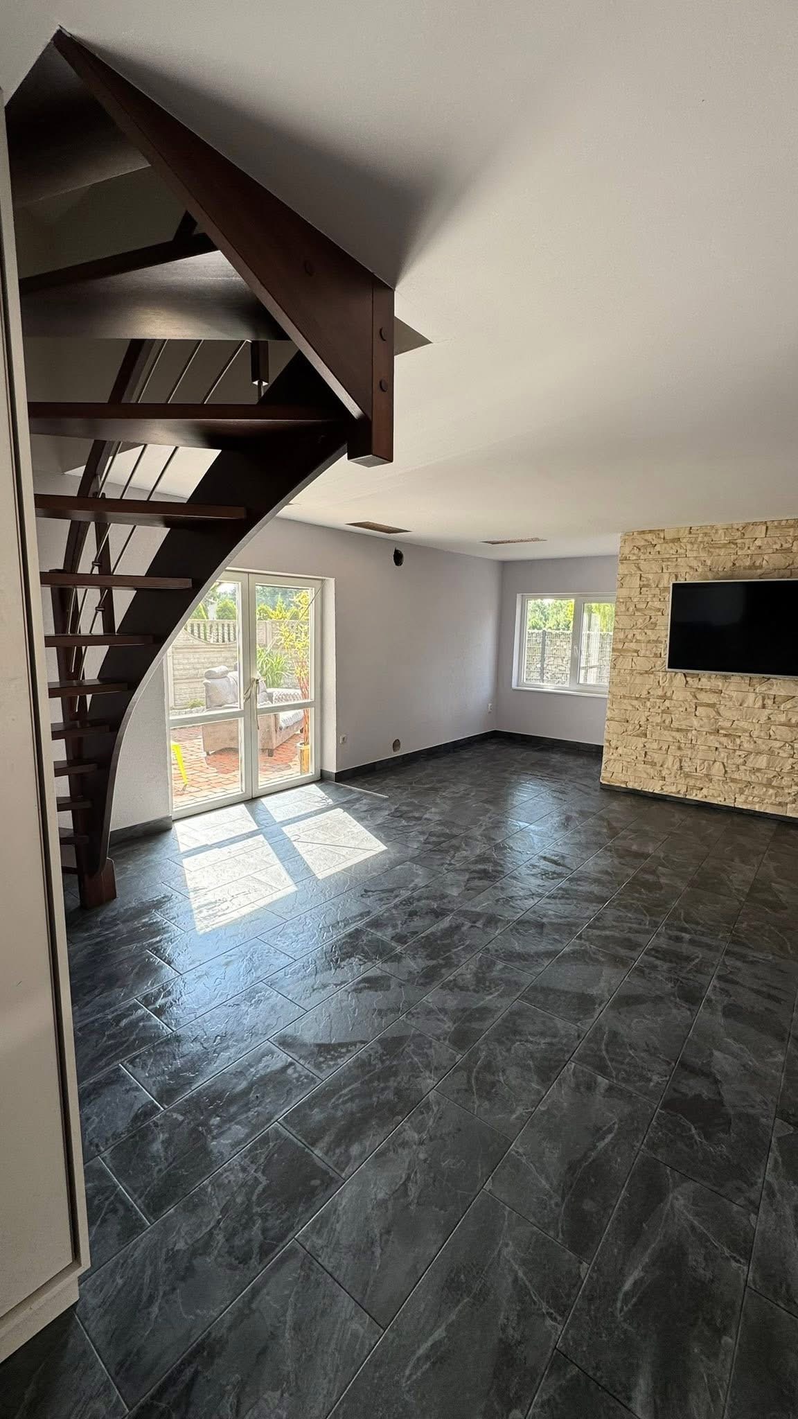 Salon po remoncie z ciemną, błyszczącą podłogą z płytek, drewnianymi schodami i ścianą z kamienia dekoracyjnego pod telewizorem. Wpadające światło słoneczne.