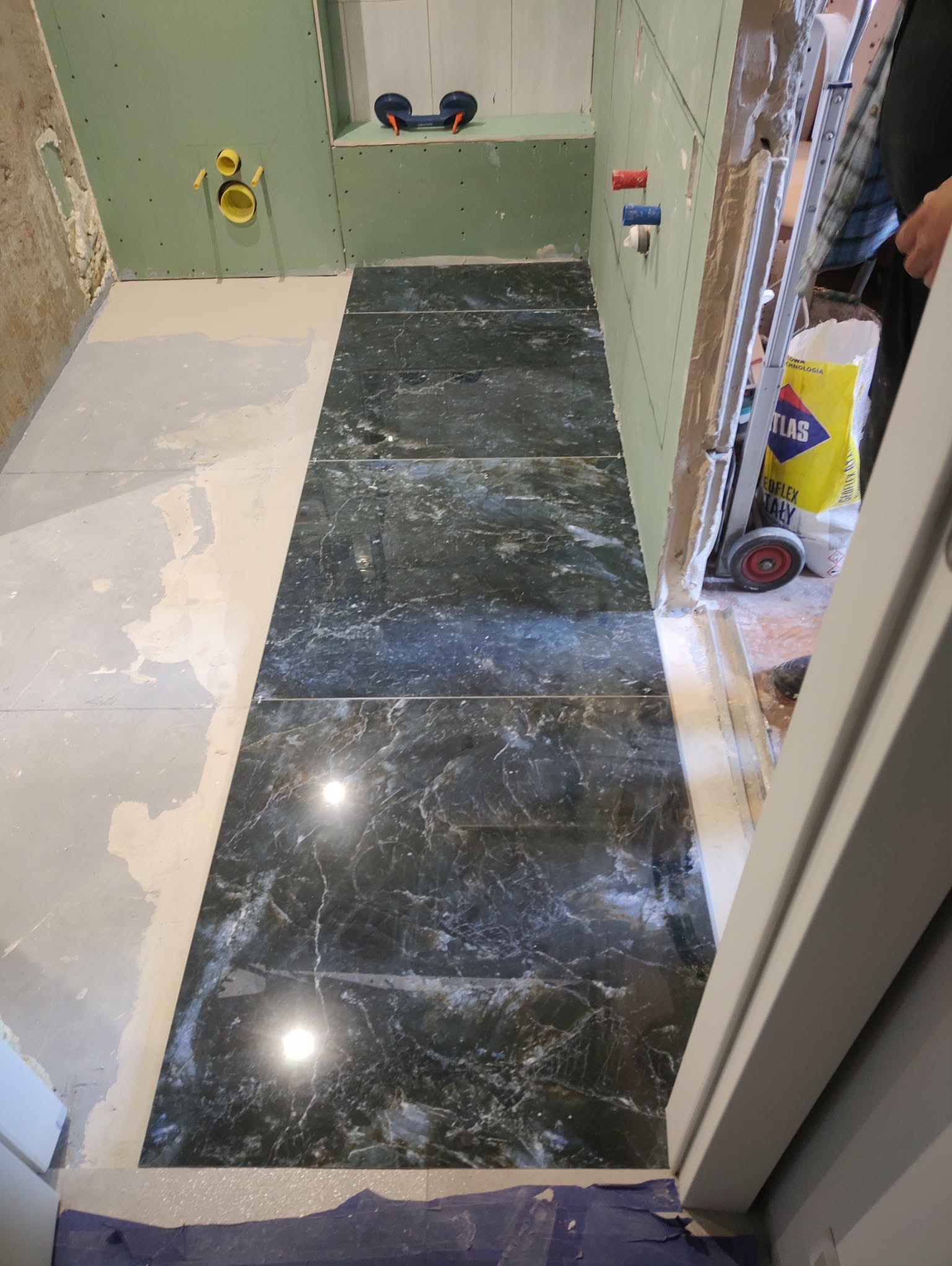 Układanie płytek podłogowych w łazience z zielonymi płytami gipsowymi na ścianach. Widoczny worek z klejem Atlas i przyssawka do płytek.