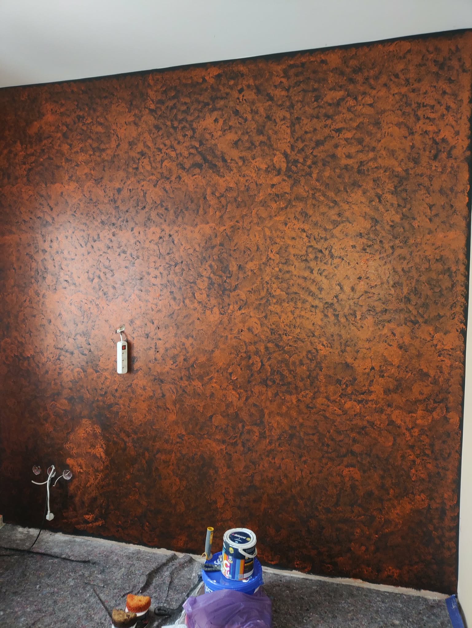 Ściana z dekoracyjną, pomarańczowo-brązową strukturą, widoczne puszki z farbą i zabezpieczenie podłogi. Gniazdka elektryczne wystają ze ściany.