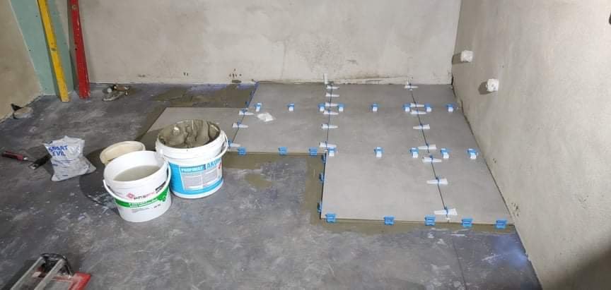 Układanie płytek podłogowych w łazience, widoczne poziomice, wiaderka z zaprawą i płytki z krzyżykami dystansowymi, surowe ściany w tle.