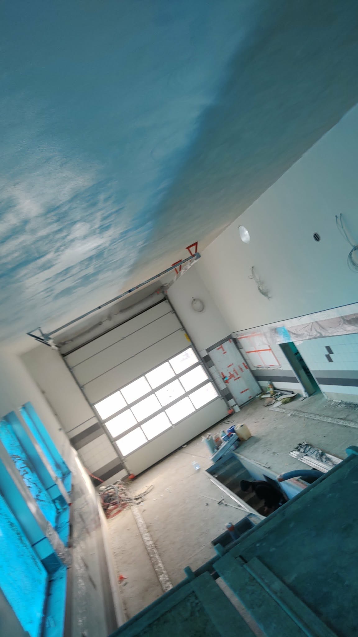 Wnętrze garażu w trakcie prac wykończeniowych: pomalowany sufit z efektem chmur, brama segmentowa, widoczne materiały budowlane i instalacje.