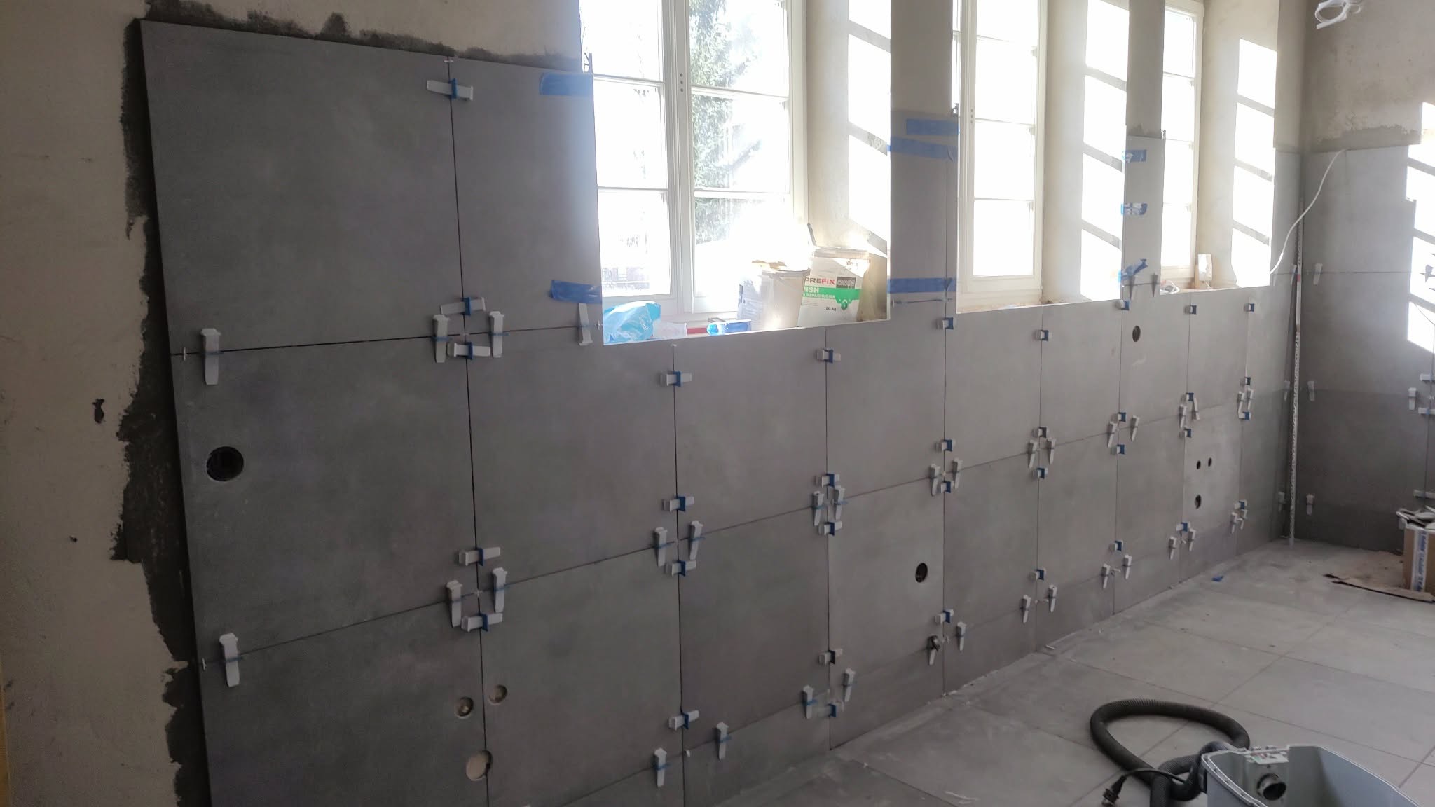 Ściana z szarymi płytkami w trakcie układania, widoczne krzyżyki dystansowe i taśma maskująca przy oknie. Częściowo ułożona podłoga i narzędzia w tle.