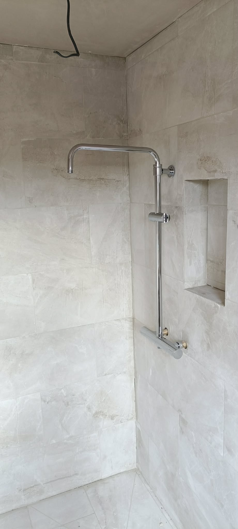 Wykończona łazienka z chromowaną armaturą prysznicową i wnęką w ścianie wyłożonej marmurkową płytką. Widoczny przewód elektryczny w rogu.