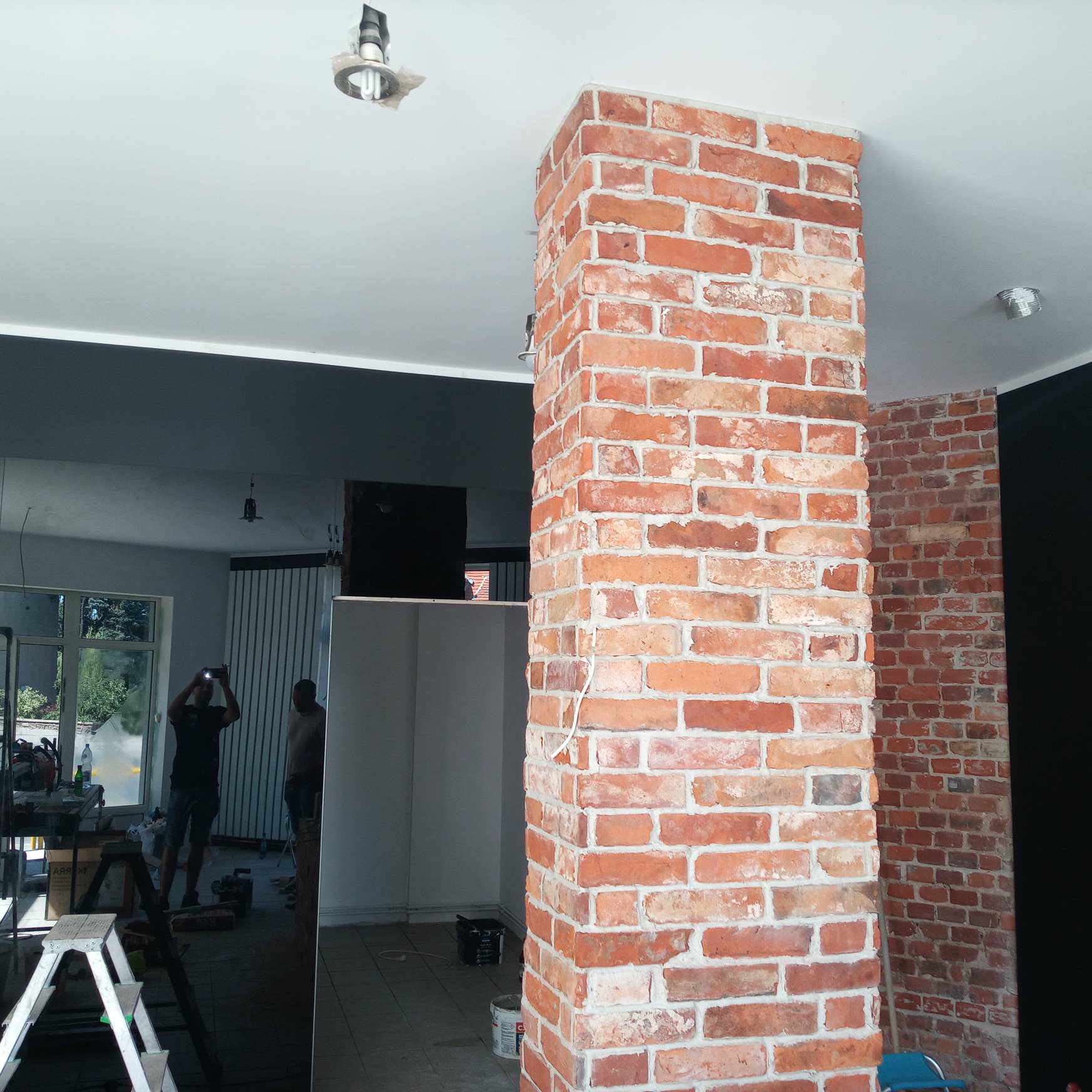 Ceglana kolumna w odcieniach czerwieni i bieli, stanowiąca element dekoracyjny wnętrza. Widoczne fragmenty pomieszczenia w trakcie prac wykończeniowych.