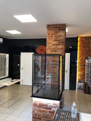 Wnętrze salonu optycznego z ceglanym filarem, czarnymi ścianami i ekspozycją okularów. Widoczna witryna i stolik z akcesoriami. Oświetlenie sufitowe.