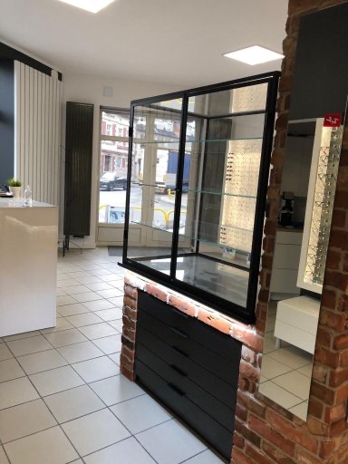 Nowoczesna witryna sklepowa z czarną ramą i szklanymi półkami, wbudowana w ścianę z cegły, z czarnymi szufladami u dołu. Widok na ulicę przez szybę.