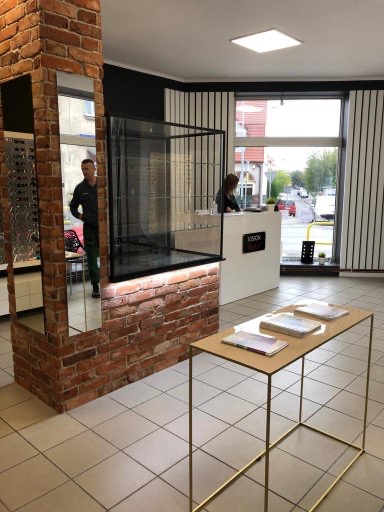 Nowoczesne wnętrze salonu optycznego z ceglaną ścianą, przeszkloną witryną i minimalistycznym stolikiem ekspozycyjnym. Widoczna recepcja i osoba za ladą.