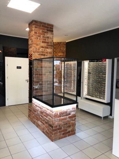 Wnętrze sklepu optycznego z ceglanym filarem i witryną, oświetleniem LED, białymi meblami i szarą ścianą w tle. Nowoczesny design z elementami industrialnymi.