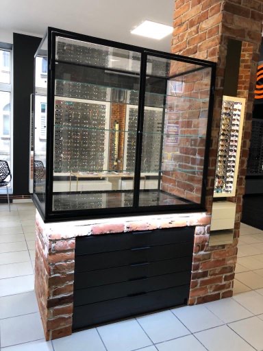 Witryna sklepowa z czarną ramą i szklanymi półkami, umieszczona na postumencie z czerwonej cegły i czarnymi szufladami, eksponująca okulary w salonie optycznym.