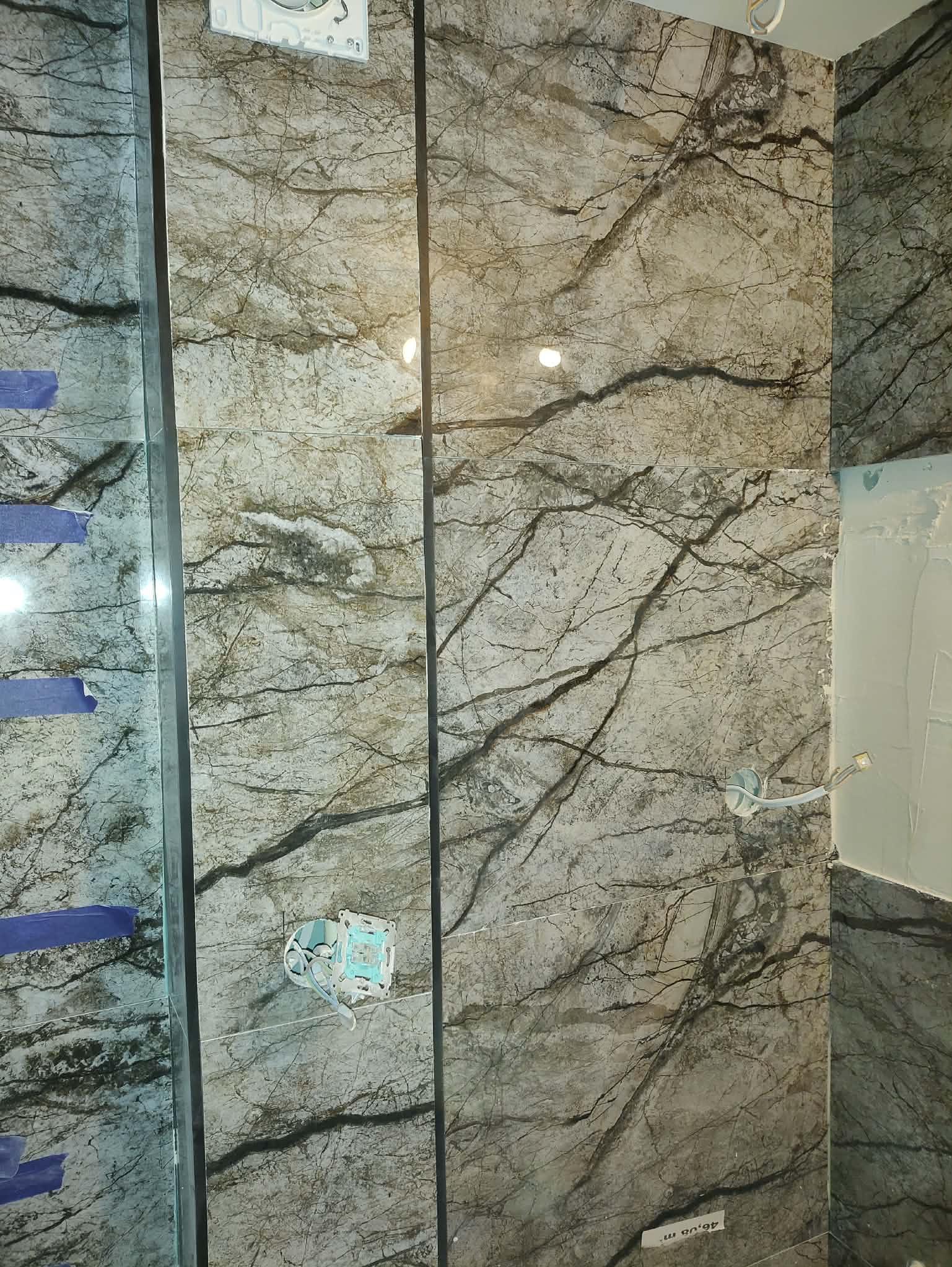 Ściana wyłożona płytkami imitującymi marmur z widocznymi puszkami elektrycznymi. Fragment lustra z niebieską taśmą zabezpieczającą. Wykończenie łazienki w trakcie.