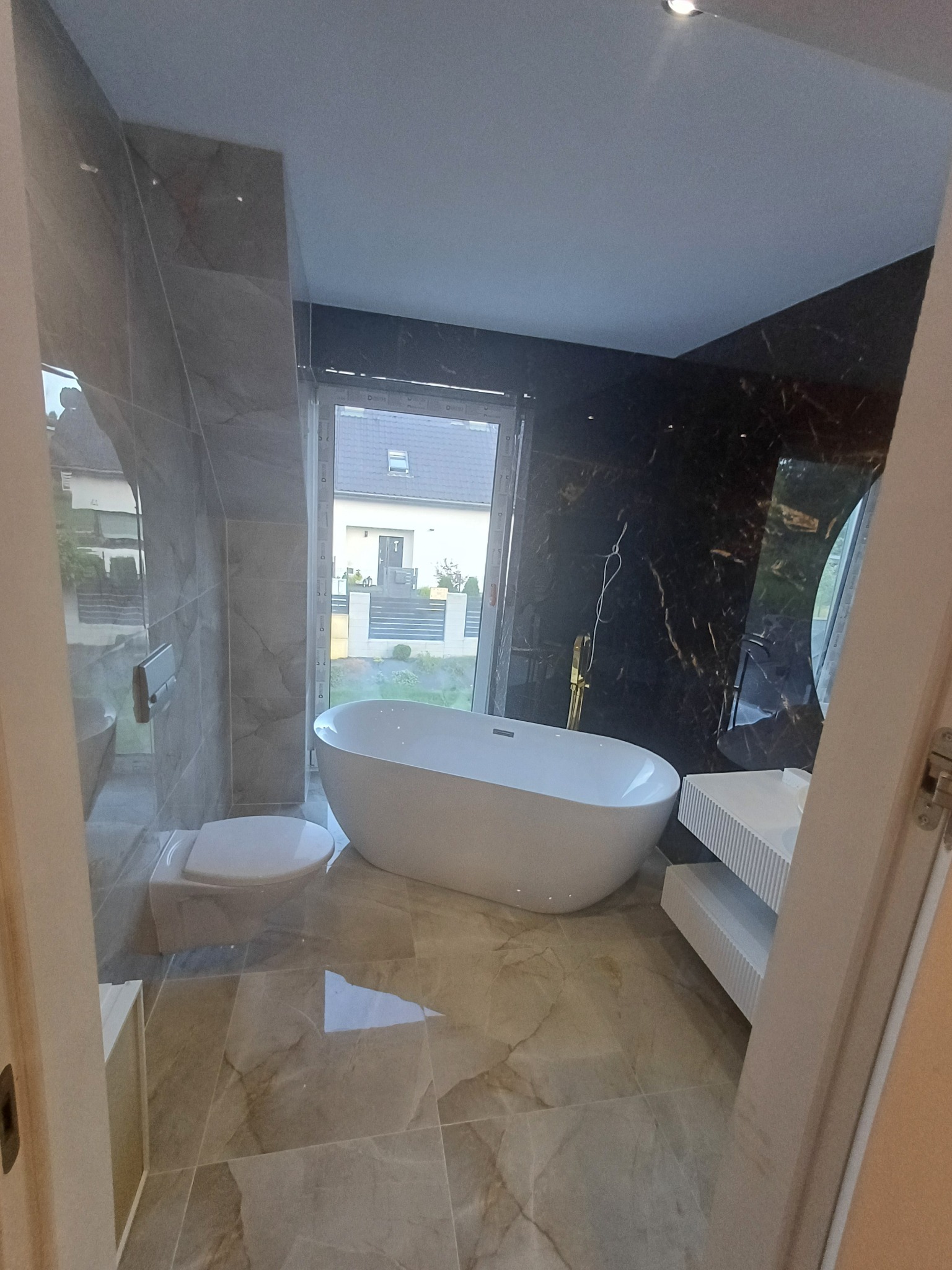 Elegancka łazienka z wolnostojącą wanną, marmurowymi płytkami i oknem z widokiem na dom. Nowoczesny design z elementami złota i bieli.