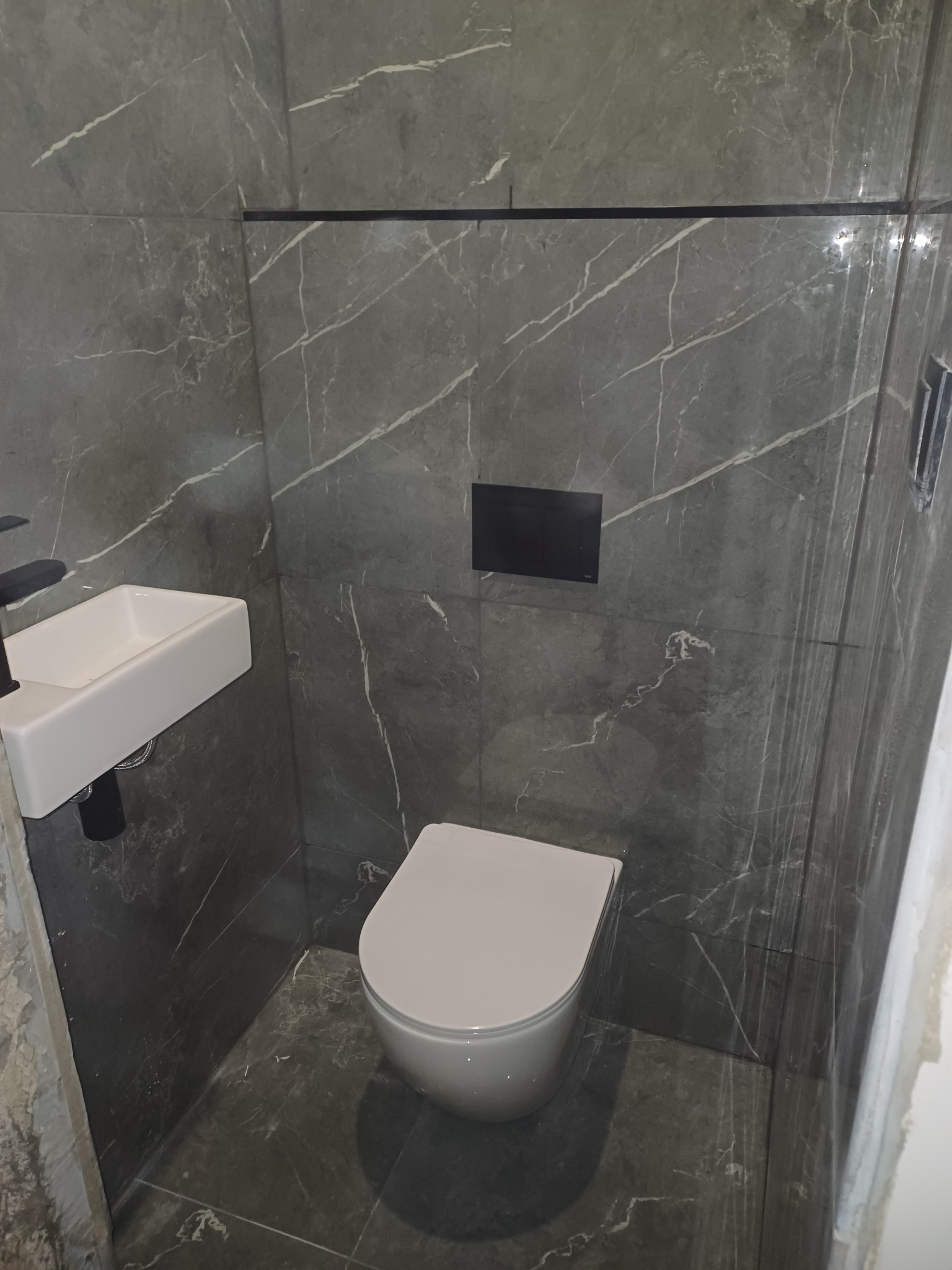 Nowoczesna toaleta z szarymi płytkami imitującymi marmur, podwieszana miska WC i umywalka. Minimalistyczny design, czarne akcenty.