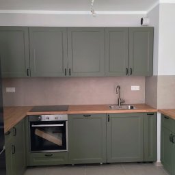 Złota Śrubka Kacper Szymczyk - Kompletna zabudowa kuchenna w odcieniach zieleni z drewnianym blatem, obejmująca szafki wiszące i stojące, piekarnik oraz lodówkę. Nowoczesny design z minimalistycznymi uchwytami.