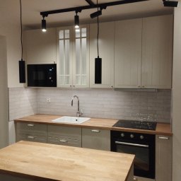 Złota Śrubka Kacper Szymczyk - Jasna kuchnia z drewnianym blatem i wyspą. Białe płytki na ścianie, nowoczesne oświetlenie. Ustawienie mebli w kształcie litery L. Widok na piekarnik i zlew.