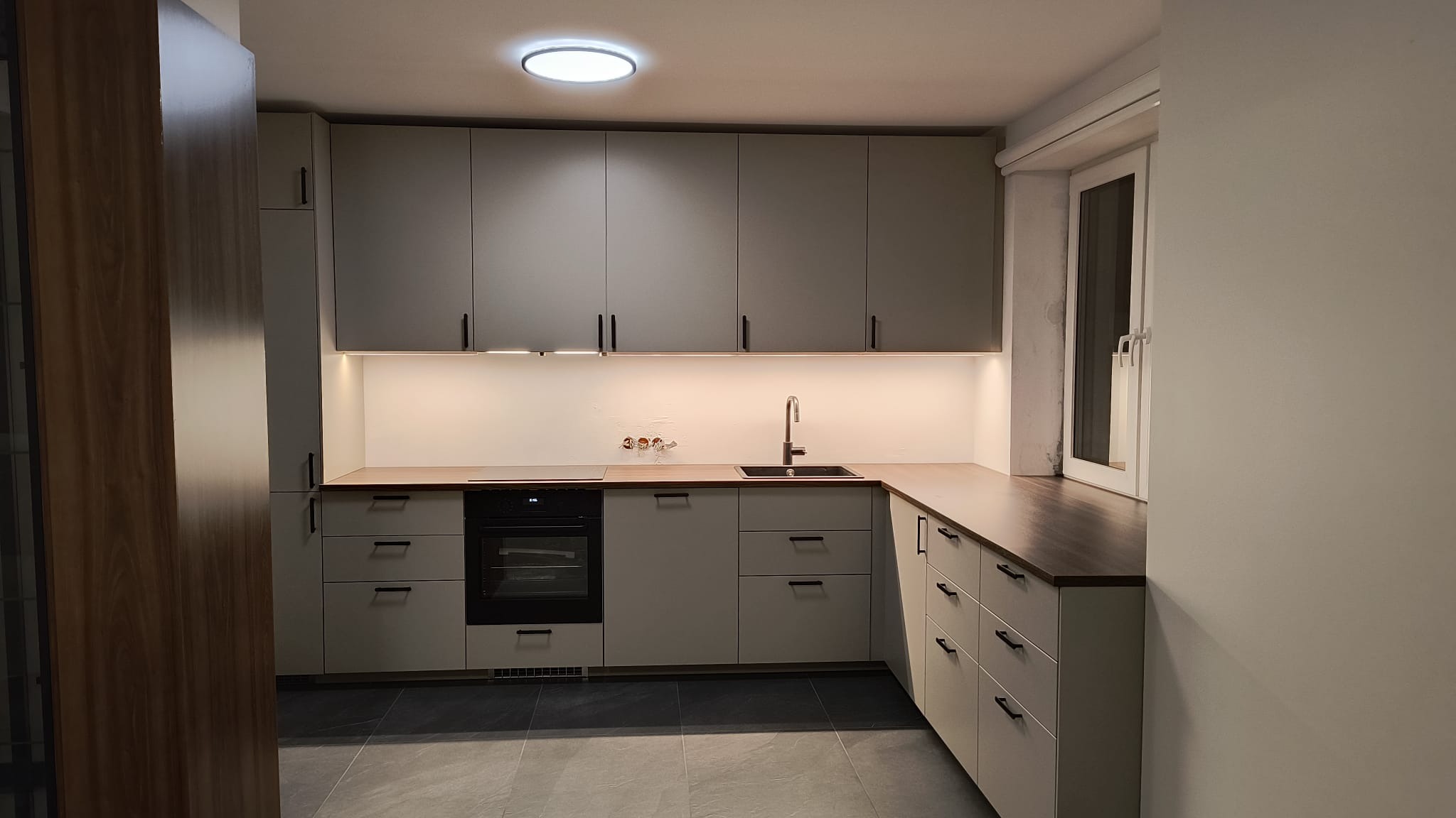 Nowoczesna kuchnia z szarymi szafkami, drewnianym blatem i oświetleniem LED pod szafkami wiszącymi. Widoczne okno i ciemna podłoga. Minimalistyczny design.