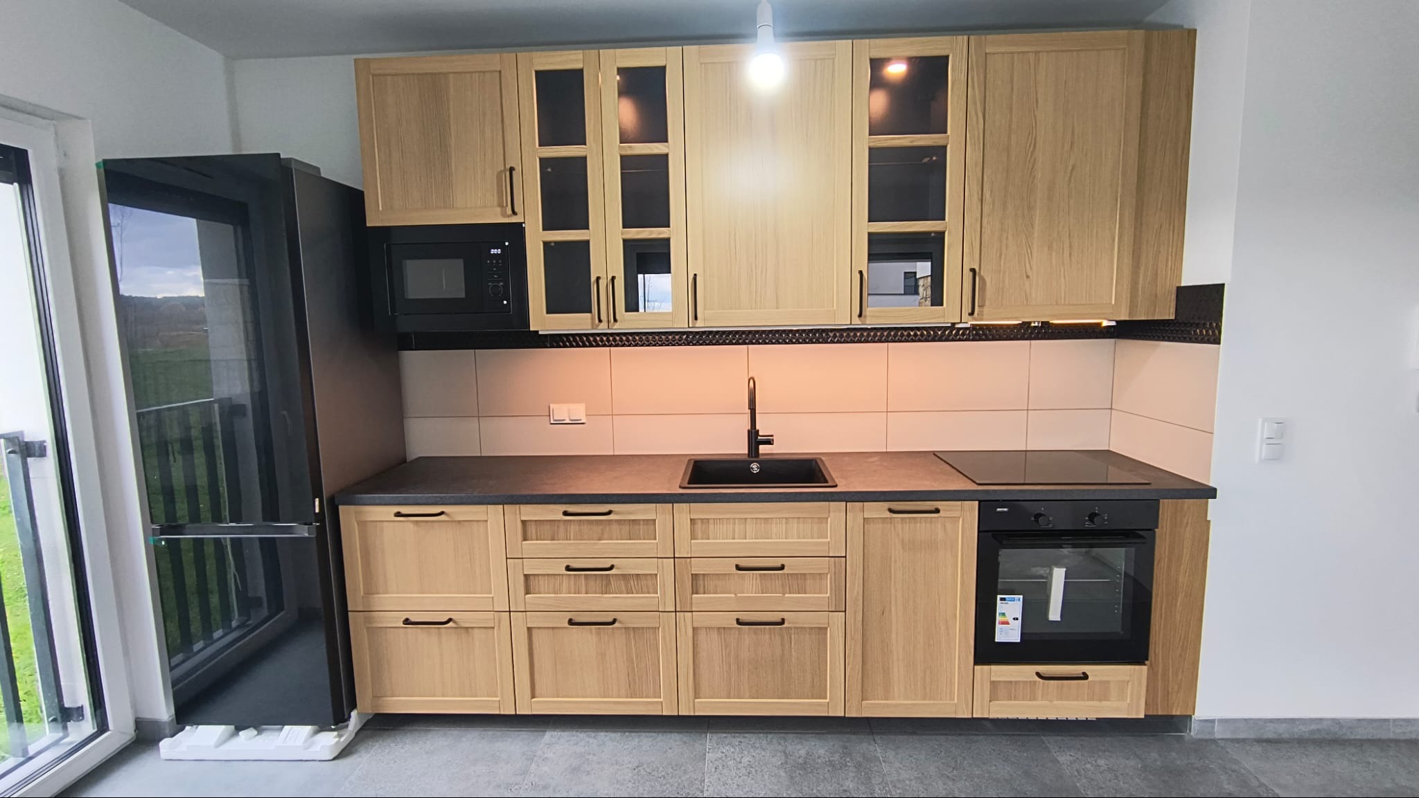 Jasna, drewniana zabudowa kuchenna z czarnymi akcentami: zlew, piekarnik, mikrofalówka i lodówka. Nowoczesny design, oświetlenie podszafkowe, minimalistyczny styl.