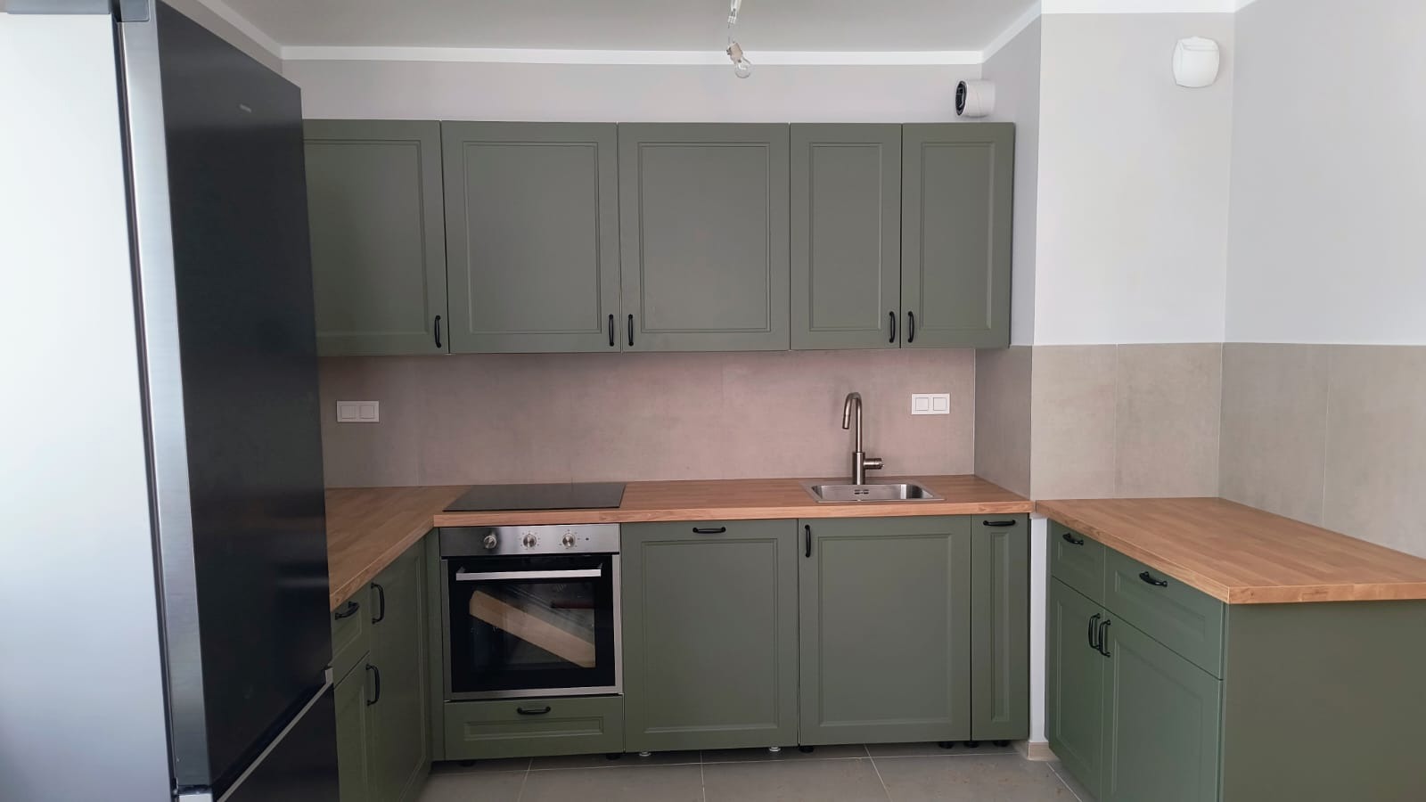 Kompletna zabudowa kuchenna w odcieniach zieleni z drewnianym blatem, obejmująca szafki wiszące i stojące, piekarnik oraz lodówkę. Nowoczesny design z minimalistycznymi uchwytami.