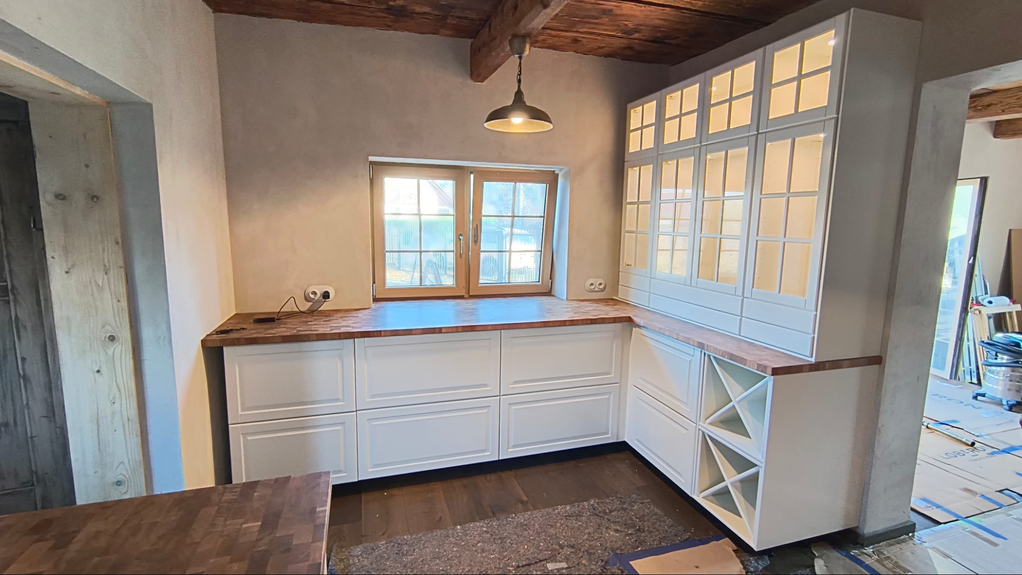 Biała zabudowa kuchenna z drewnianym blatem i oświetloną witryną, widok na okno i sufit z belkami. Nowoczesny design w rustykalnym wnętrzu.