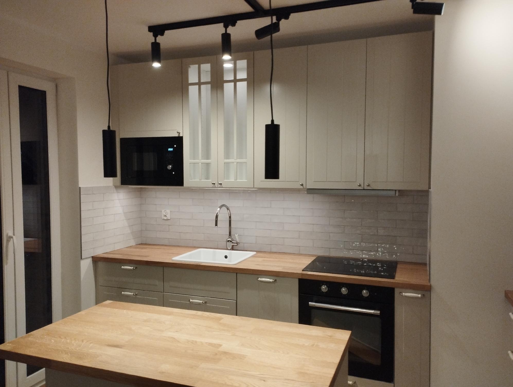 Jasna kuchnia z drewnianym blatem i wyspą. Białe płytki na ścianie, nowoczesne oświetlenie. Ustawienie mebli w kształcie litery L. Widok na piekarnik i zlew.