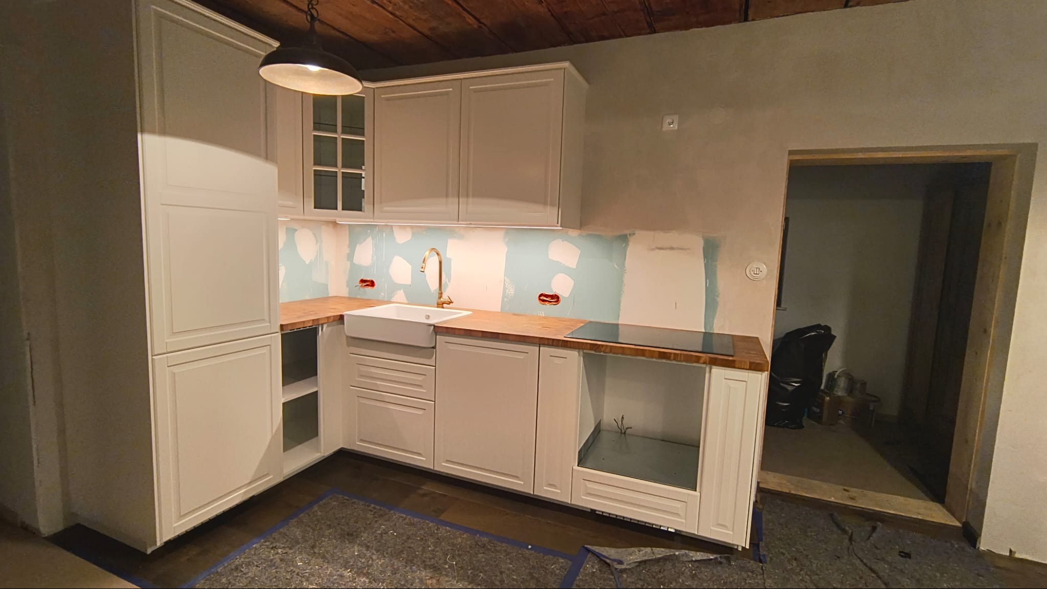 Jasna kuchnia z drewnianym blatem i złotą armaturą, w trakcie prac wykończeniowych. Widoczne ślady po malowaniu na ścianach i surowy drewniany sufit.