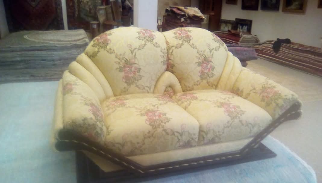 Dwuosobowa sofa tapicerowana w kremowy materiał z motywem różowych kwiatów, na tle dywanów i obrazów. Widoczne detale wykończenia i charakterystyczny kształt mebla.