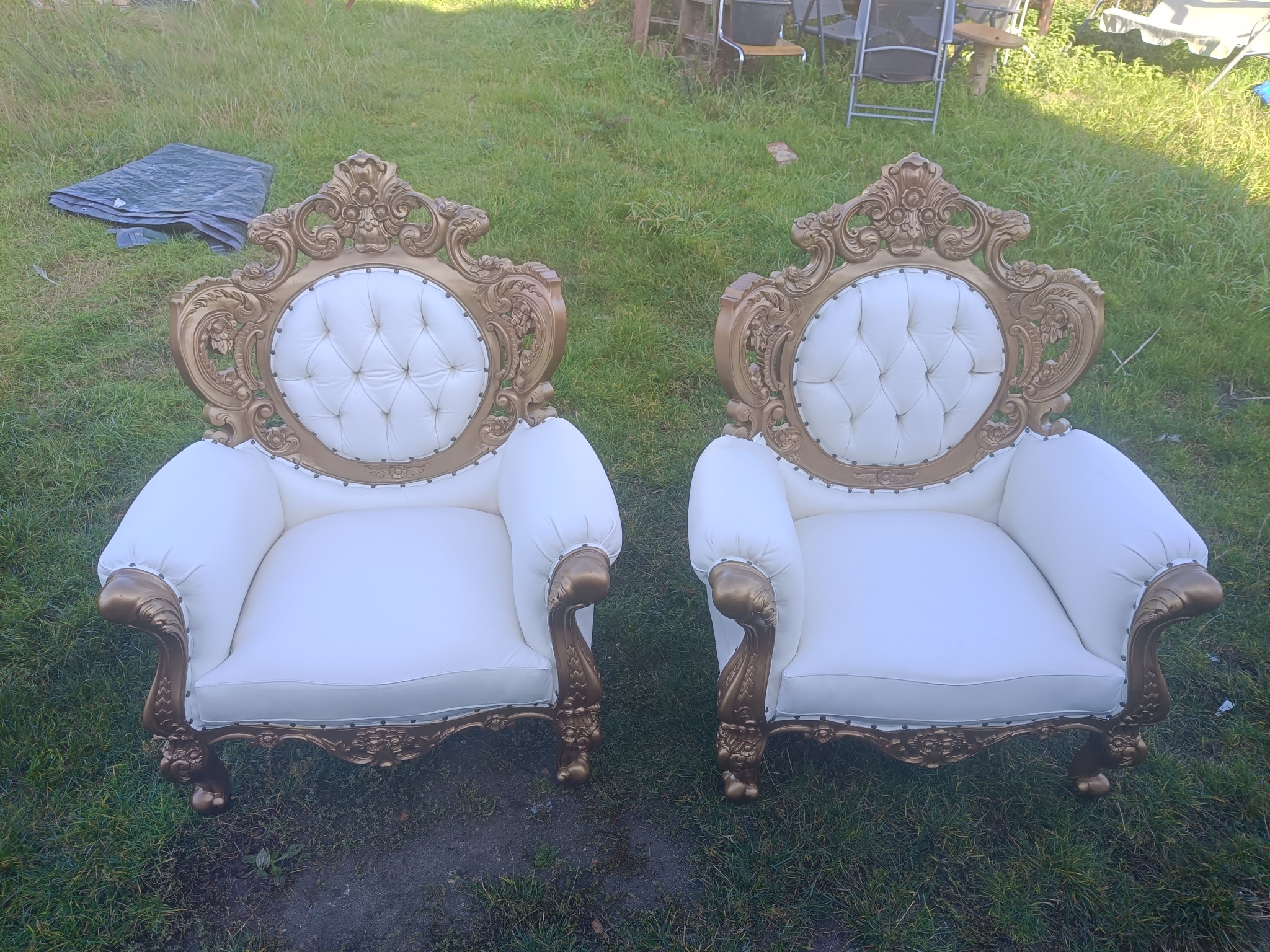 Dwa identyczne, odnowione fotele w stylu barokowym z białą tapicerką i złotymi zdobieniami, ustawione na trawie. Widoczne detale rzeźbione w drewnie.