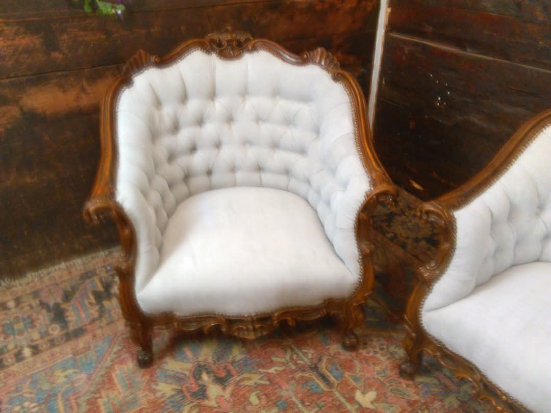 Antyczny fotel z jasną tapicerką i rzeźbionymi, drewnianymi elementami, ustawiony na wzorzystym dywanie. Widoczny fragment drugiego fotela.