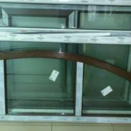 Dwa białe ramy okienne z szybami, dolna z łukowatym brązowym elementem dekoracyjnym, ułożone jedno na drugim na podłodze z płytek.