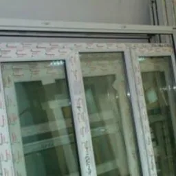 Stos białych, nie zamontowanych ram okiennych z szybami, widoczne profile z nadrukowanym wzorem, ustawione obok siebie.