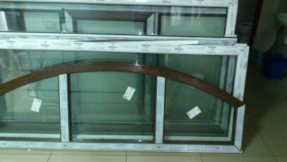 Dwa białe ramy okienne z szybami, dolna z łukowatym brązowym elementem dekoracyjnym, ułożone jedno na drugim na podłodze z płytek.